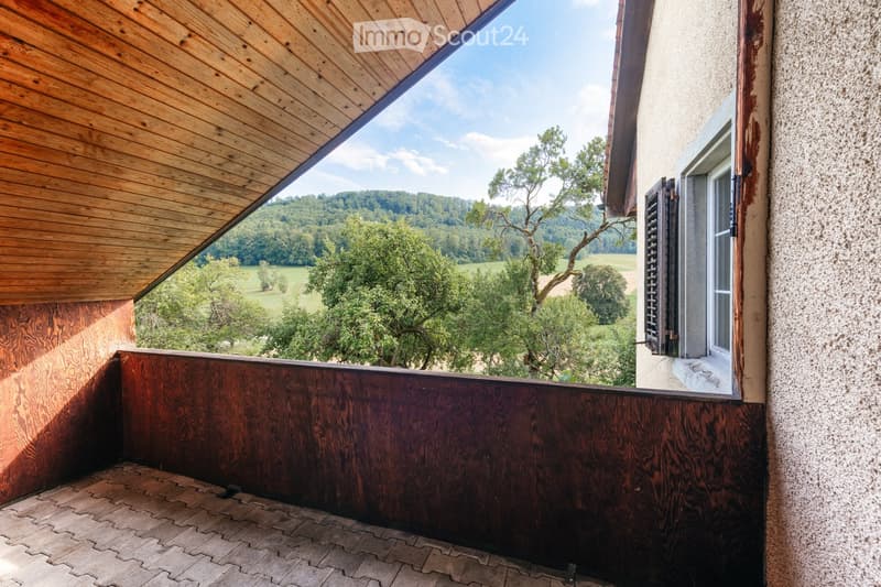 Wooden balcony with a slanted roof, overlooking a green landscape with trees and fields.