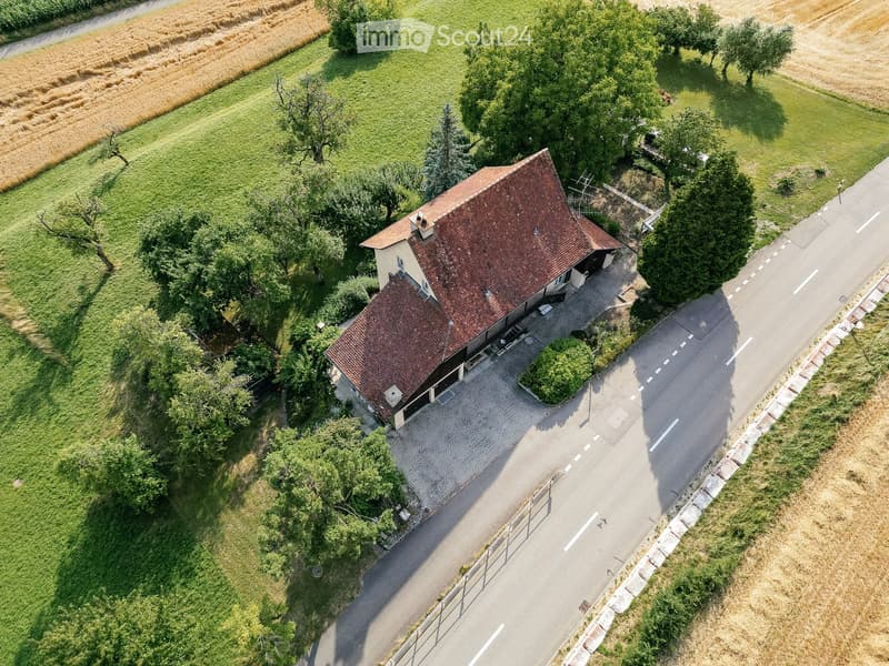 Aerial view of a two-story house with a red-tiled roof, surrounded by a paved driveway and greenery. The house is situated near a road with a grassy field and trees in the background.