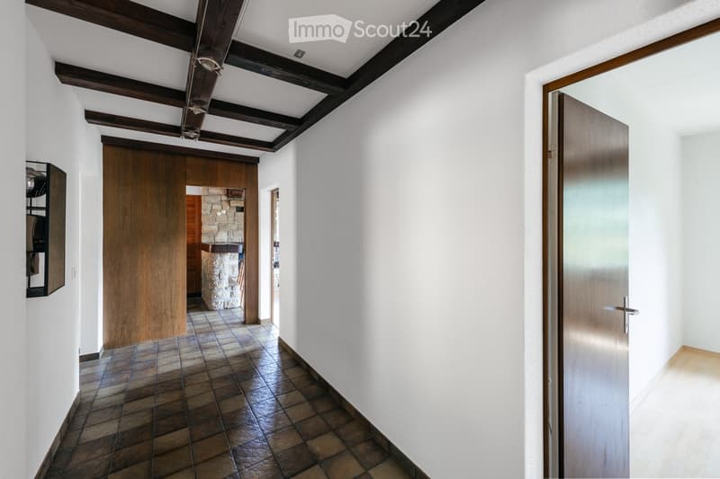 A spacious hallway with exposed wooden beams on the ceiling, tiled floor, and white walls. Features include a wooden door on the right, a stone pillar in the background, and a small black shelf on the left wall.