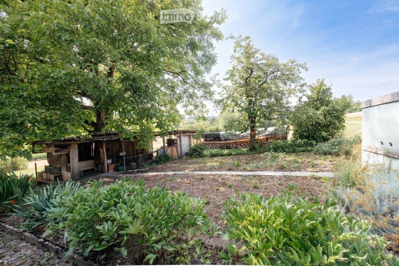 A serene garden area with a wooden shed, a stone wall, and a variety of plants and trees. The garden is bordered by a gravel path and features a mix of greenery and open space.