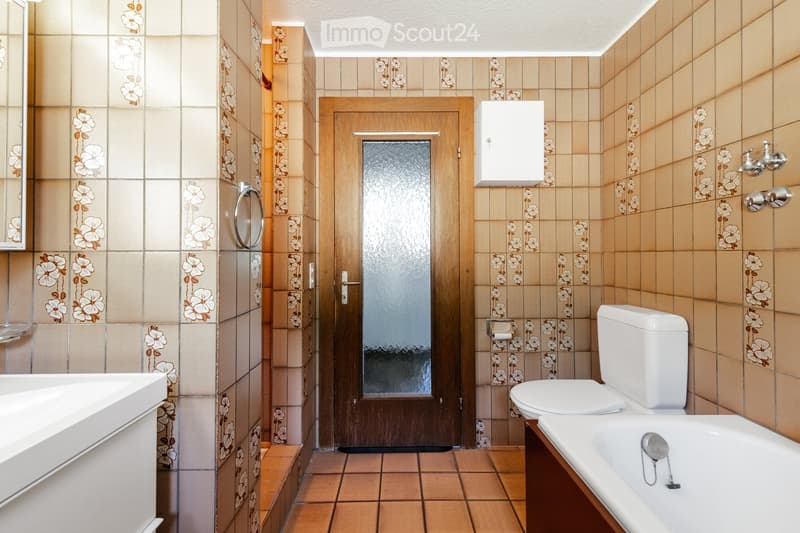 Bathroom with floral-patterned tiles, a toilet, bathtub, sink, and a wooden door with a frosted glass panel.