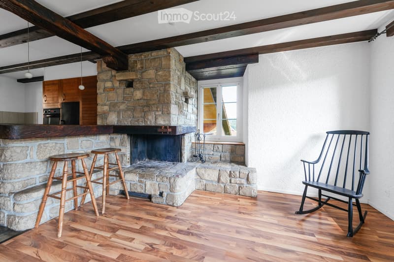 Living room with exposed wooden beams, stone fireplace, wooden stools, rocking chair, and a small kitchen area visible in the background.