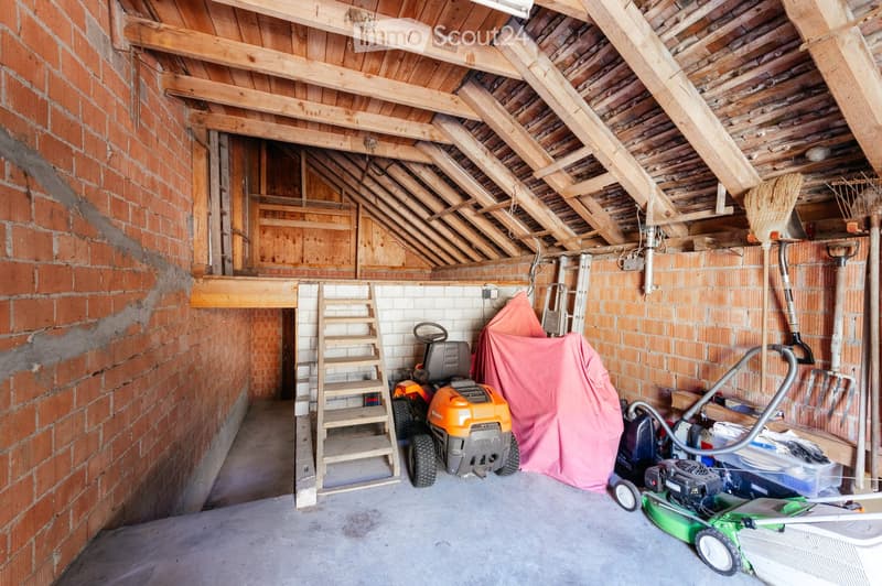Garage with exposed wooden beams, brick walls, and various stored items including lawn mowers, a ladder, and a broom.