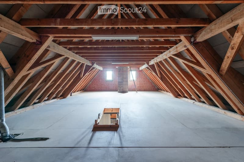 An empty attic space with exposed wooden beams and rafters, concrete floor, and small windows allowing natural light.
