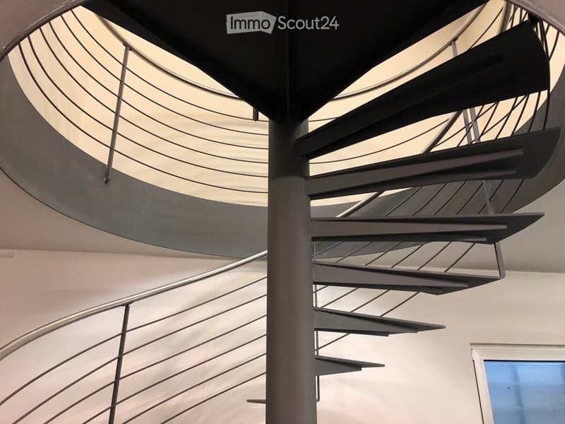 Modern, curved staircase with metal railings and steps, featuring a central column and a glass ceiling allowing natural light to filter through.