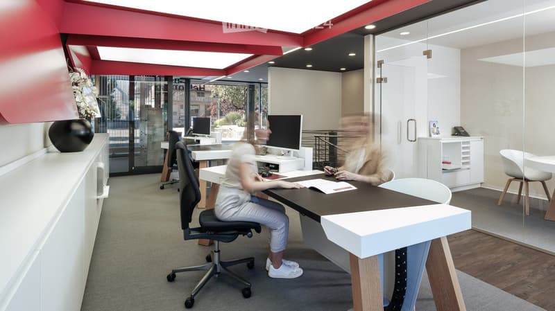 Modern office space with a black table, white chairs, and a computer screen. The room features a sleek design with red and white walls, large windows, and a vase on a cabinet.