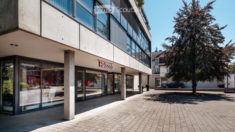 Modern building entrance with glass doors and large windows, featuring the 'Helsana' logo, surrounded by a paved area with trees and a parked car.