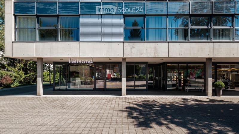 Modern building entrance with large glass windows and doors, concrete columns, and a paved area.