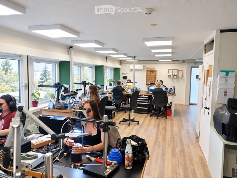 A modern office space with multiple workstations, each equipped with microscopes, laptops, and various tools. Employees are seated at desks, working with equipment. The room features large windows with a view of greenery outside, wooden flooring, and brig