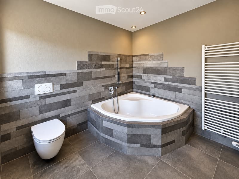 Modern bathroom with a corner bathtub, toilet, grey tiled walls, and a white radiator.