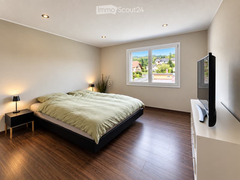 Modern bedroom with wooden flooring, large window with scenic view, bed with beige bedding, nightstand with lamp, television on stand, and decorative plant in corner.