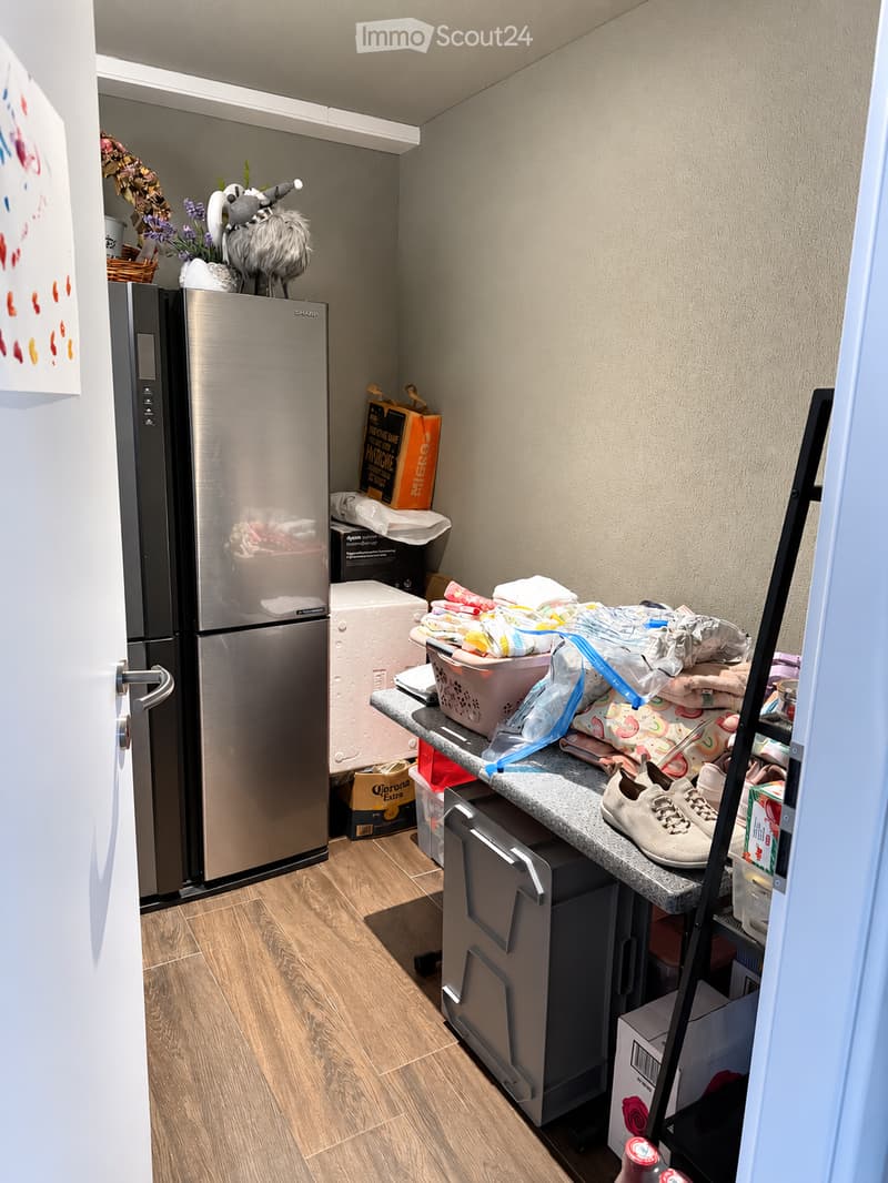 A small, furnished room with a refrigerator, microwave, counter, and various items including a basket, shoes, and boxes.