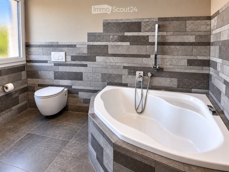 Modern bathroom with a large bathtub, toilet, and sleek grey tiles. Features a handheld shower hose and a window with frosted glass.