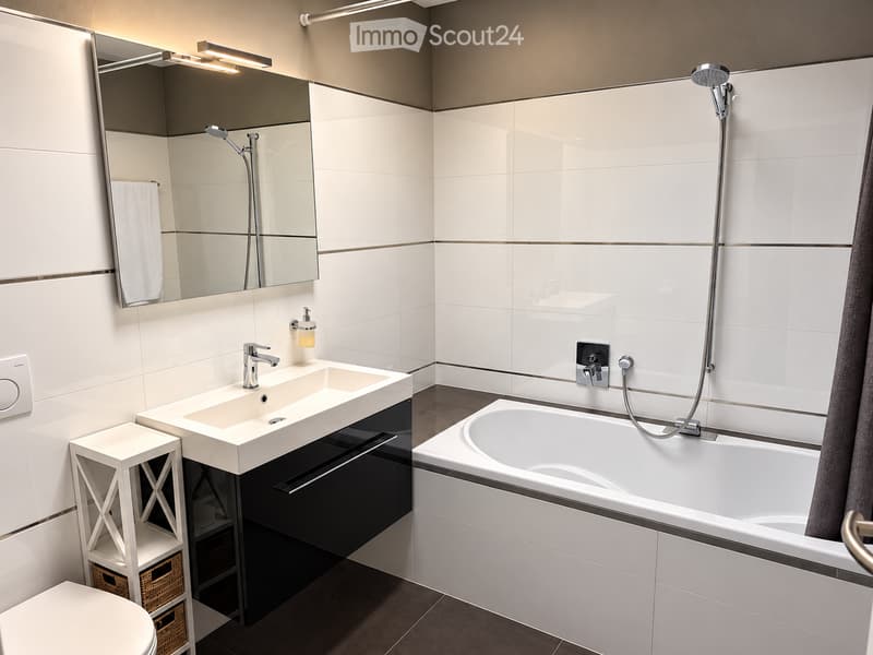 Modern bathroom with white tiles, a large mirror, a sink with a modern faucet, a bathtub with a handheld showerhead, and a small white stool with woven baskets underneath.