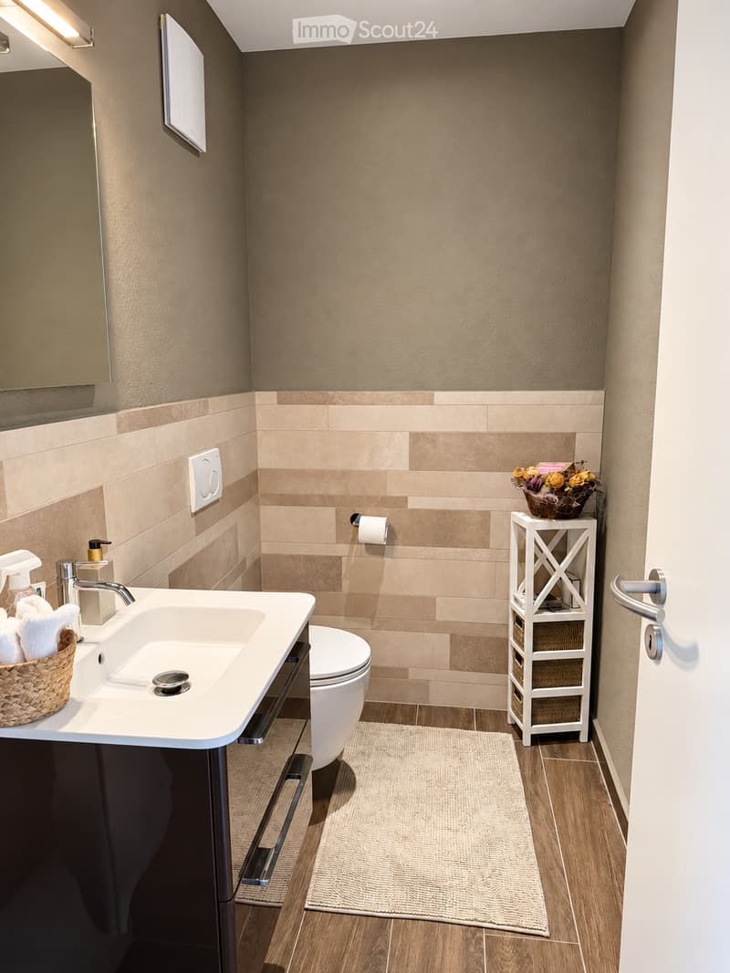 Modern bathroom with beige walls, wooden flooring, a white sink with a chrome faucet, a toilet, a towel basket, a small shelf with a flower arrangement, and a mirror above the sink.