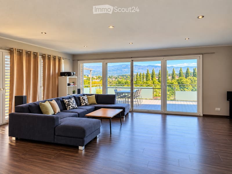 Modern living room with dark blue sectional sofa, wooden coffee table, large sliding glass doors leading to a balcony with outdoor furniture and scenic mountain view.