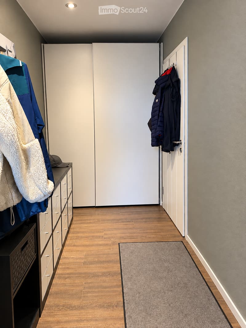 Clean, modern hallway with wooden flooring, neutral-colored walls, and built-in storage units on one side. A small rug is placed in the center, and coats are hanging on hooks near the door.