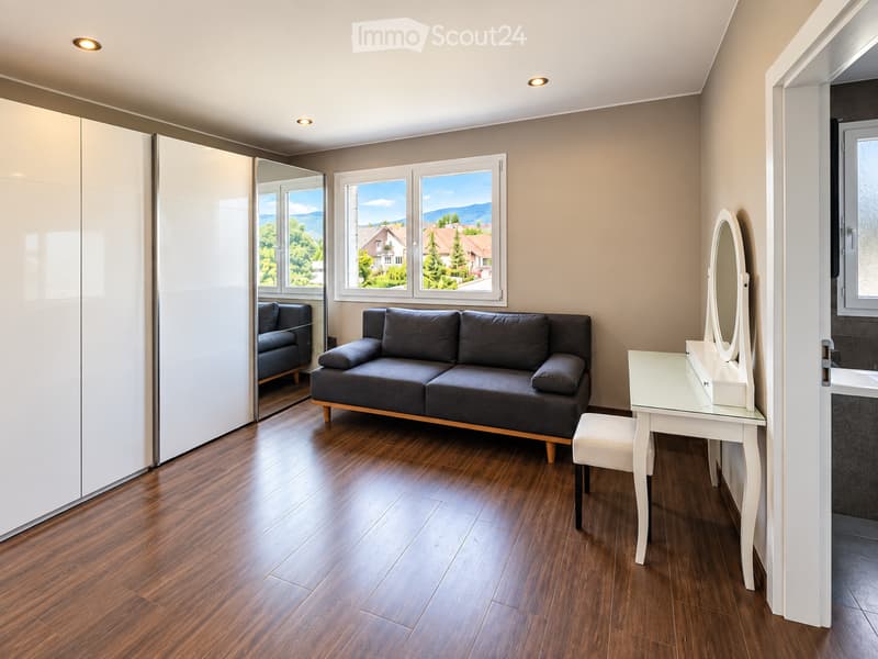 Modern living room with wooden flooring, large windows offering a scenic view, a grey sofa, a white vanity table with a mirror, and a white wardrobe.