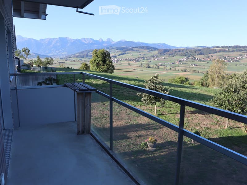 Balcony with metal railing, overlooking a scenic valley and distant mountains, with a small wooden bench and potted plant visible.