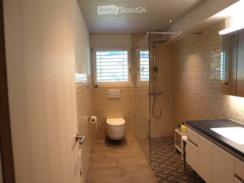Modern bathroom with beige tiles, a glass-enclosed shower, a toilet, a sink with a mirror, and a small cabinet holding toiletries.