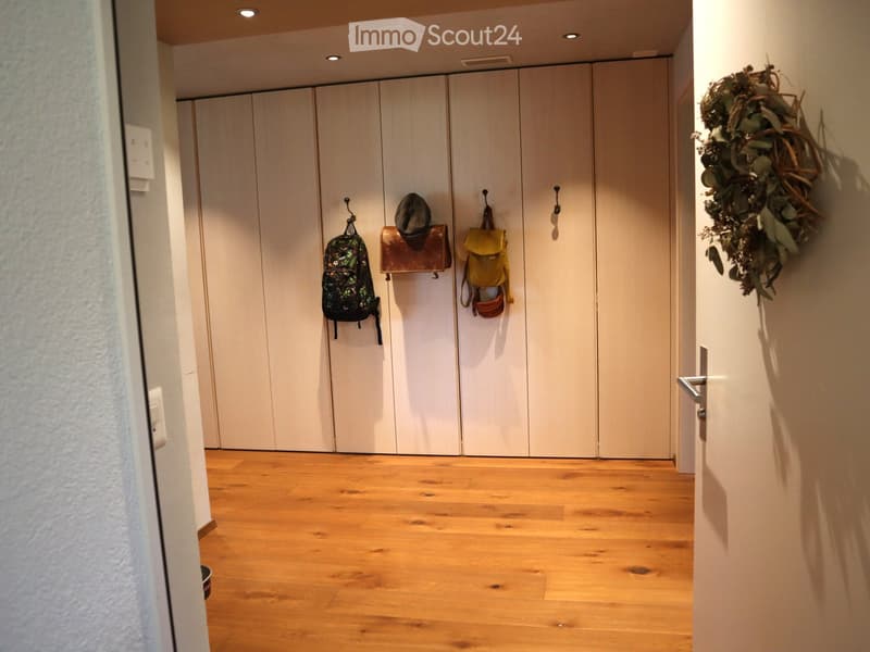 A modern hallway with light wooden flooring, white closet doors, and a few hanging items including a backpack, a hat, and a bag.