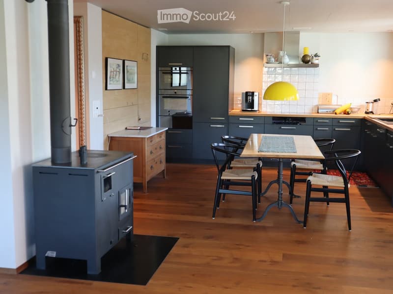 Modern kitchen with wooden flooring, grey cabinets, a central island, a dining table with chairs, and a built-in oven.