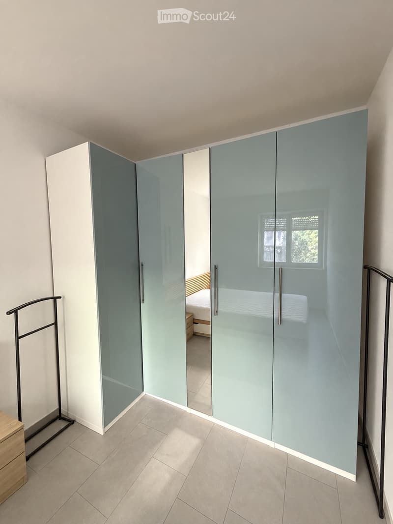 Modern bedroom with large mirrored wardrobe, bed, and nightstand visible through the open wardrobe doors.