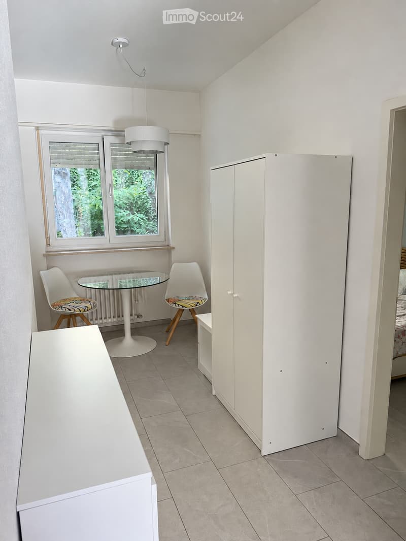 A small, modern room with a white tiled floor, a large window with blinds, a white pendant light, a round glass table with two colorful chairs, and a white cabinet.