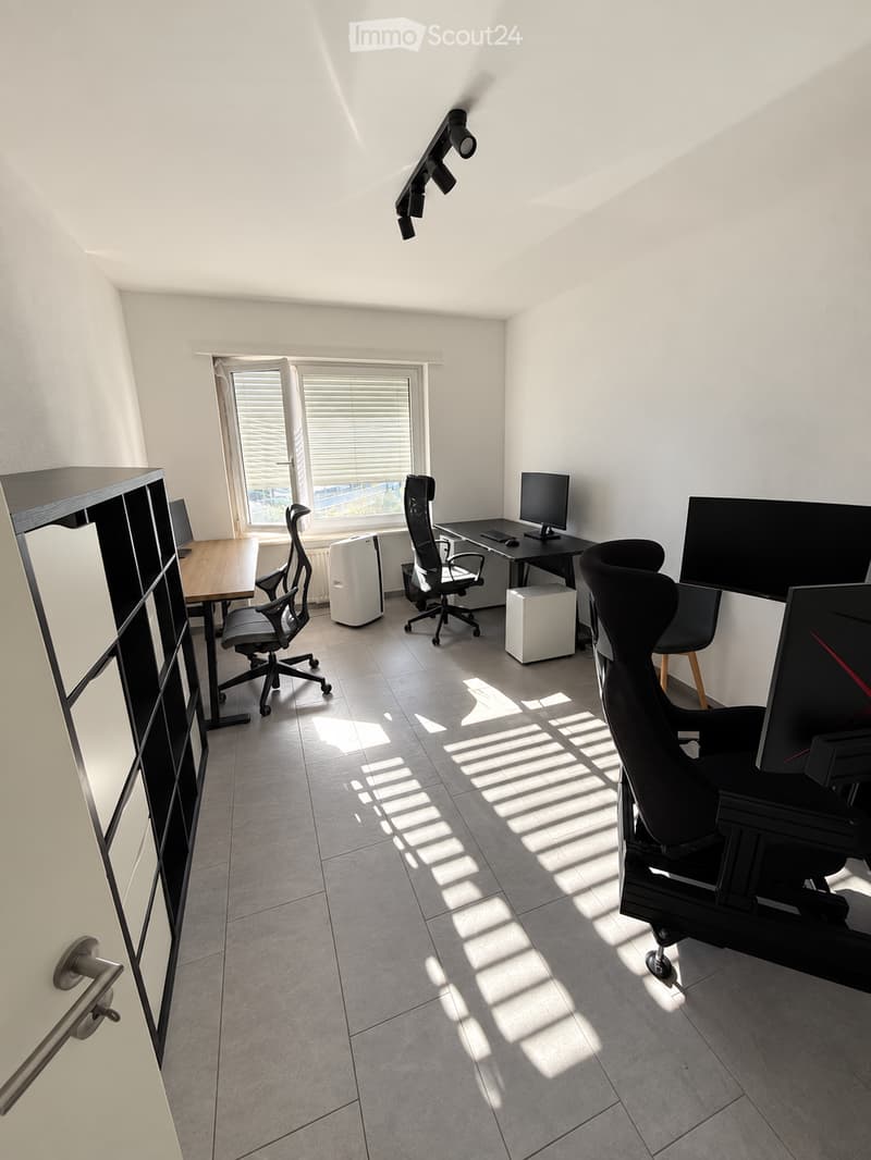 Modern office space with two desks, multiple chairs, computers, and a large window with blinds. The room features a tiled floor and white walls.