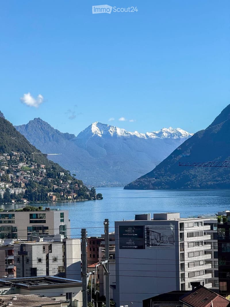 Panoramic view of a lake surrounded by mountains, with modern buildings in the foreground and a town along the lakeshore.