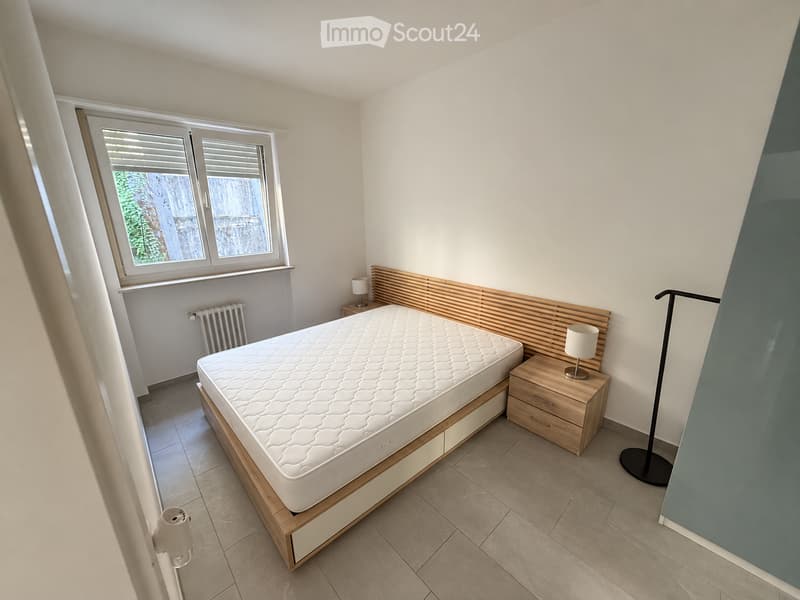 Modern bedroom with a wooden bed frame, white mattress, nightstand with lamp, radiator, and large window with blinds.