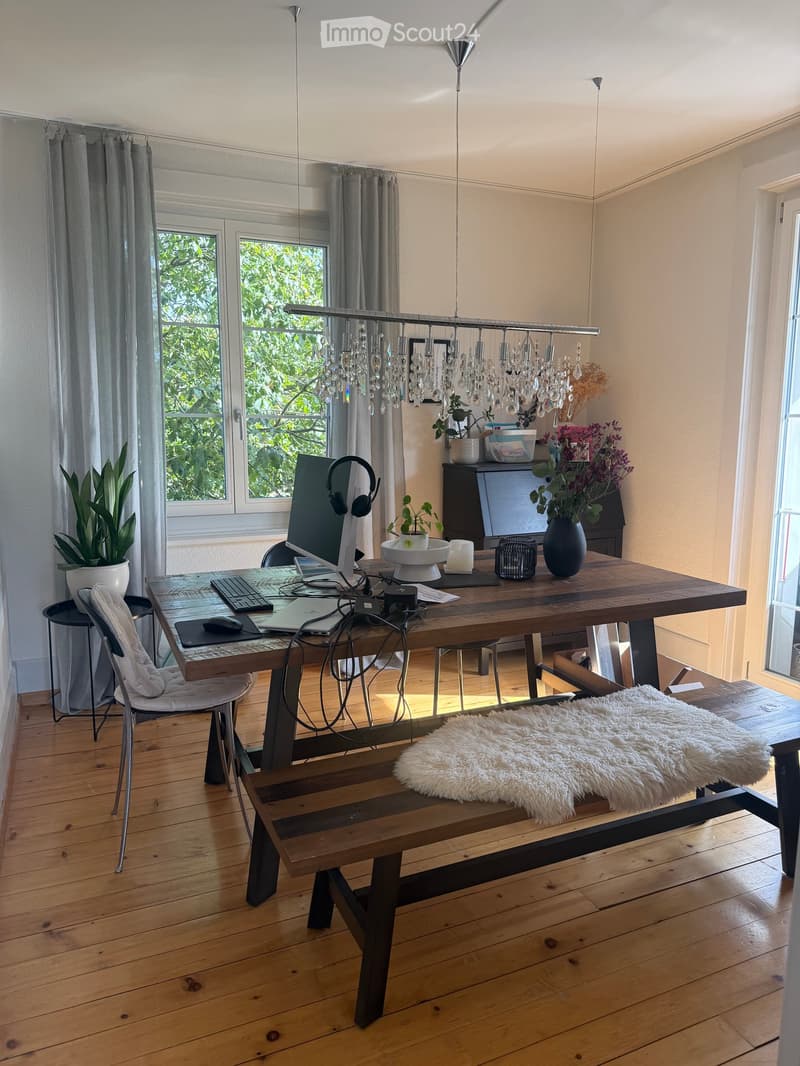 A well-lit room with wooden flooring, a large wooden table with a laptop, keyboard, mouse, and headphones, a bench with a fur throw, and a chandelier hanging from the ceiling. There are plants and a vase on the table, and large windows with curtains.