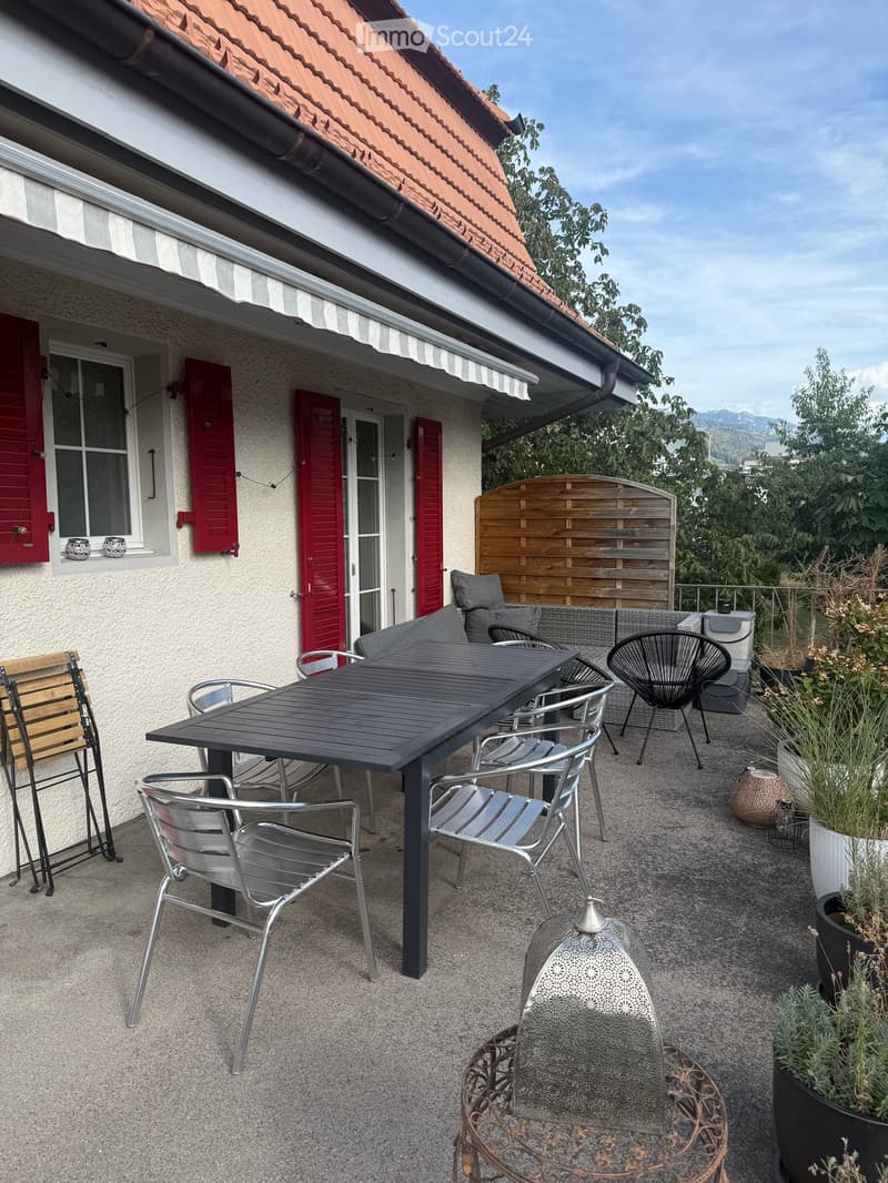 Balcony with a black table, several chairs, a sofa, and a fire pit, surrounded by potted plants and a wooden fence.