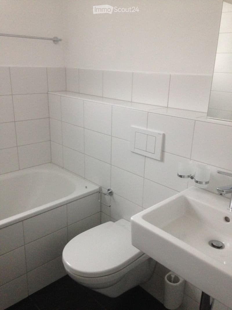 A small bathroom with white tiled walls and floor, featuring a toilet, sink, bathtub, and a mirror above the sink.