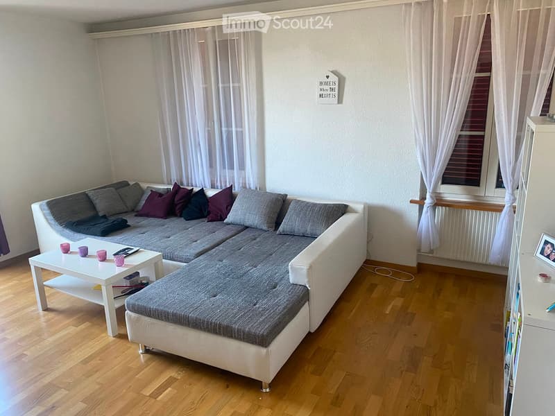 Modern living room with a grey sectional sofa, white coffee table, and decorative pillows. The room features wooden flooring, large windows with white curtains, and a small shelf with books and a picture frame.