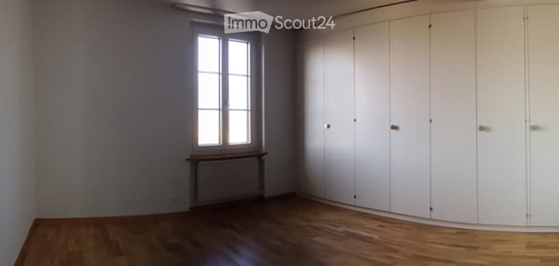 Empty room with wooden flooring, white walls, large window, built-in white closet, and a small wooden bench under the window.