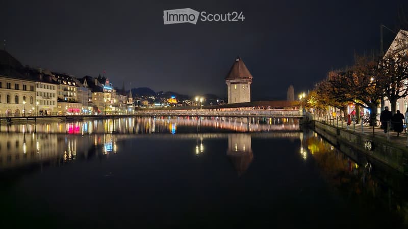 City by the river with streetlights and bridges, historic tower, buildings on both sides of the river, reflections on the water, people walking on the street.