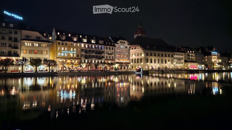 Urban setting with multistory buildings along a waterfront with lights reflecting on the water, presence of hotel signage, and historical architectural details