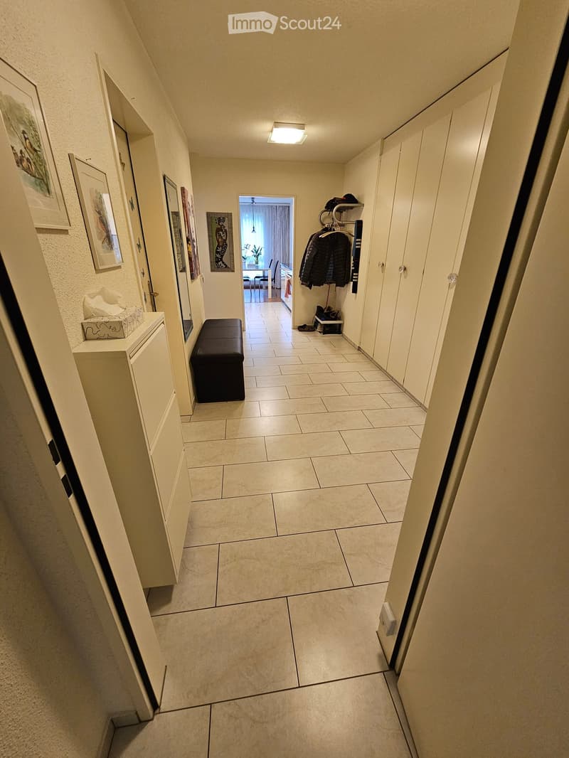 A narrow hallway with tiled flooring, white walls, and several doors. There are framed pictures on the left wall, a small bench, and a coat rack with jackets on the right side. A doorway at the end leads to another room.