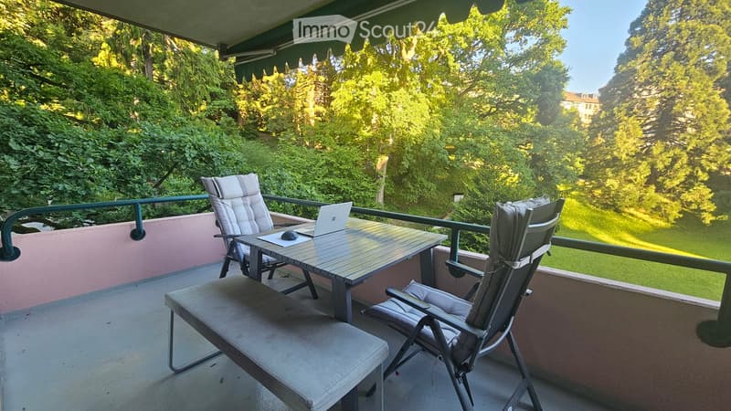 Balcony with a table, two chairs, and a laptop, overlooking a green, tree-filled area.