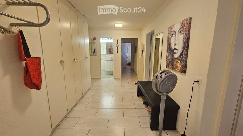 Hallway with tiled floor, white walls, and a fan placed on a small black table. A painting is visible on the right wall, and a red bag hangs on the left side.