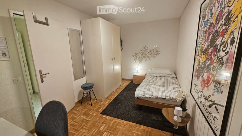 Cozy bedroom with a single bed, nightstand, small table, stool, and artwork on the wall. The room features wooden flooring and a dark rug.