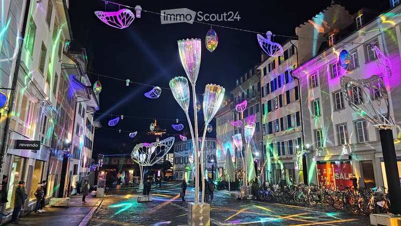 Illuminated street with shops, modern art installations, bicycles parked on the side, pedestrians walking, buildings with modern architectural designs, colorful lighting