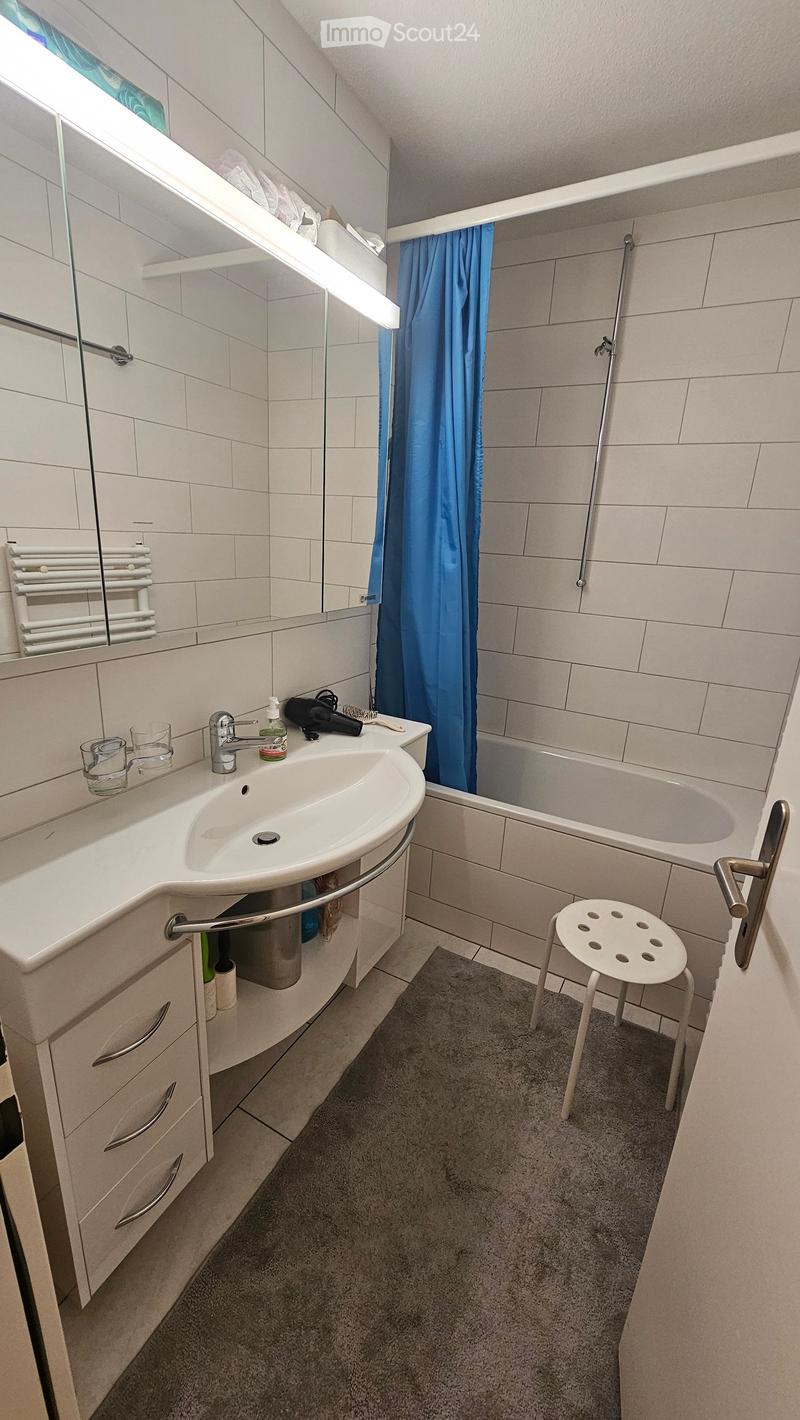 Modern bathroom with a white sink, mirror, towel rack, and a small white stool near the bathtub. The space features white tiles and a grey carpeted floor.