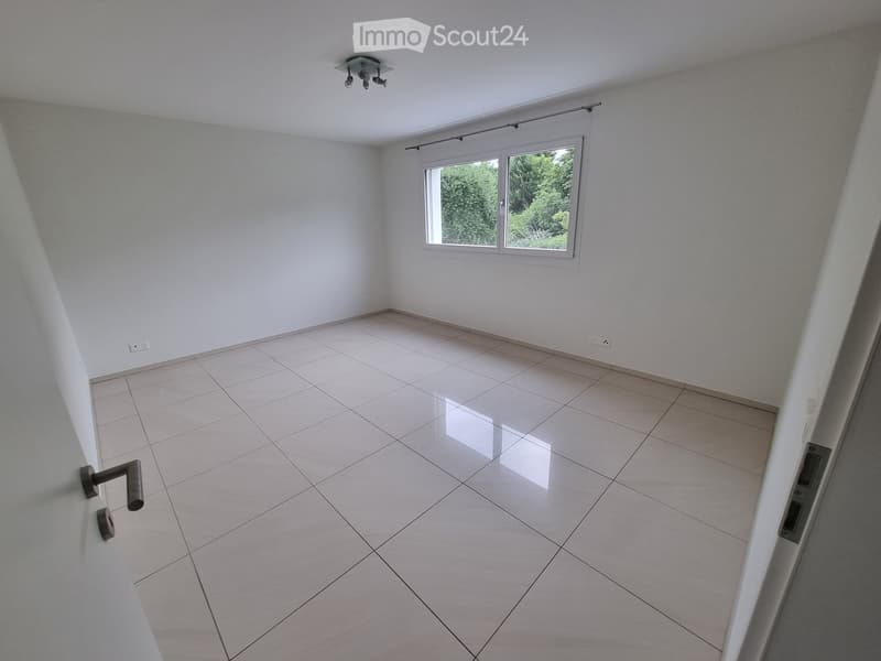 Empty room with white tiled floor, white walls, and a large window with a view of greenery. Features a ceiling light fixture and a door handle visible on the left side.