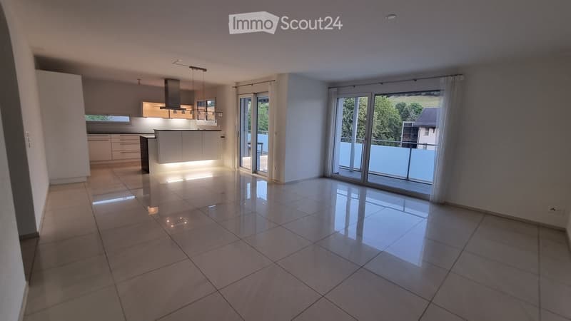 Empty room with tiled floor, large sliding glass doors leading to a balcony, and a modern kitchen area visible in the background.