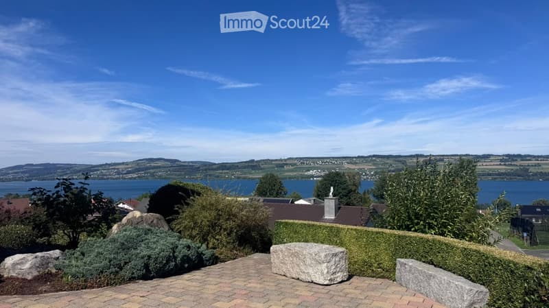 Garden with a view of a lake and surrounding hills, featuring a paved patio, stone benches, and various shrubs and trees.