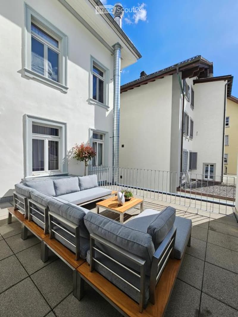patio, outdoor seating, metal railing, wooden bench, coffee table, plant in vase, white brick building, window with curtain