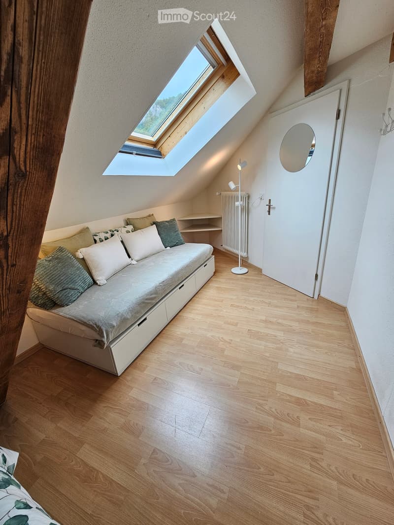 Cozy attic bedroom with wooden flooring, a built-in sofa bed, pillows, a radiator, a floor lamp, a round mirror, and built-in shelves.