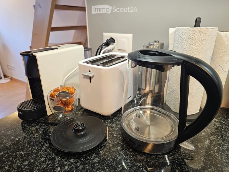 A kitchen countertop with a coffee maker, toaster, electric kettle, paper towels, a cup, and a lid.